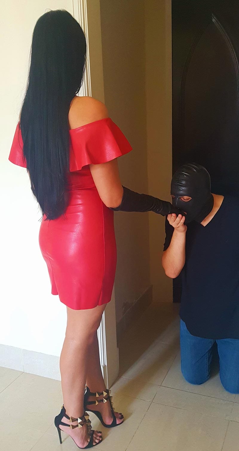 Dubai Dominatrix Anita is wearing an off the shoulders red leather dress, black designer high heels with golden details and a black leather glove which is being kissed by her slave who is kneeled in front of her.