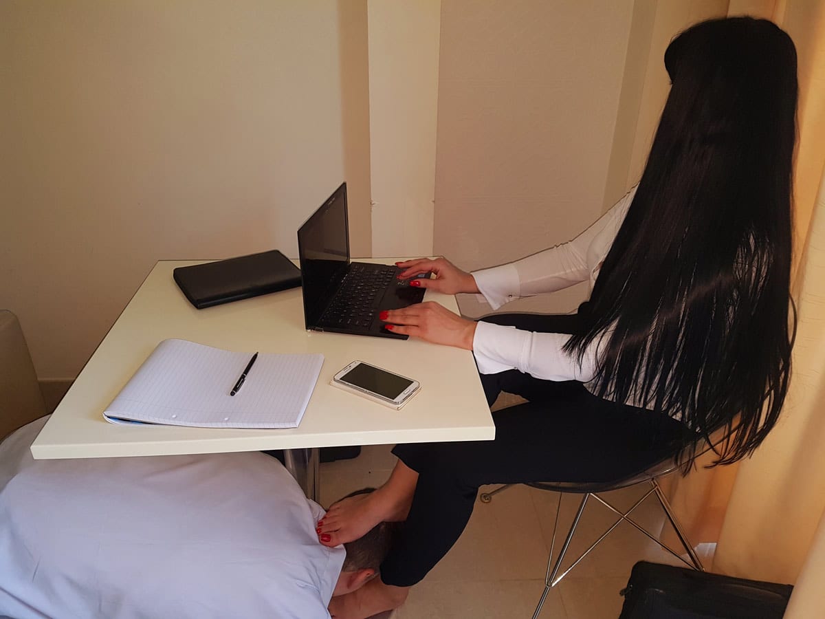 An office role play showing Mistress Anita as the C.E.O. sitting at her desk working on her Sony laptop while her sub as the badly performing employee is forced to be a foot slave to keep his job.