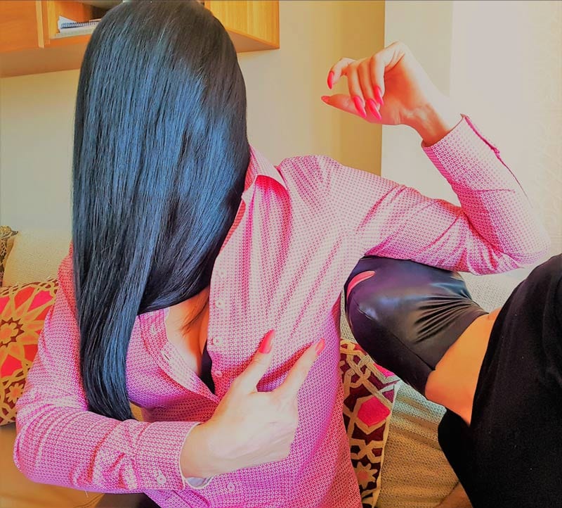 Mistress Anita is wearing a pink blouse and is pointing with her long pink nails to her armpit instructing her slave, wearing a latex mask and black t-shirt, to smell her armpit.